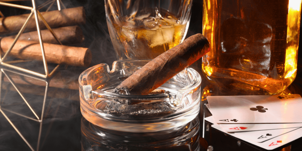 Cigar sitting in an ashtray next to a set of Ace cards and whiskey