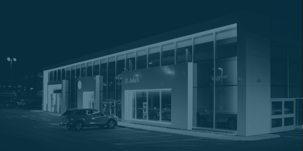 Modern car dealership building at night with illuminated showroom windows and parked vehicles outside.